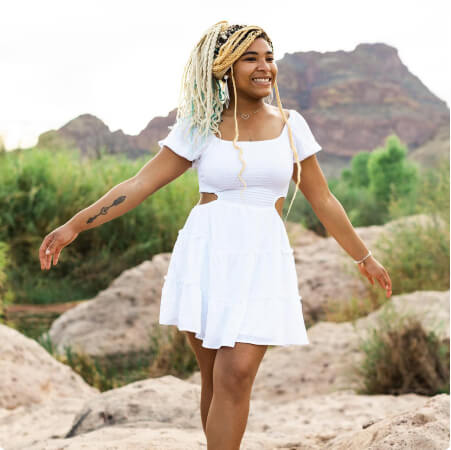 Young lady in white short sleeve, square neck dress, smocking on top, layered on bottom with open/vents on sides of waist. Standing on top of large rocks in a nature mountainous backgrouund