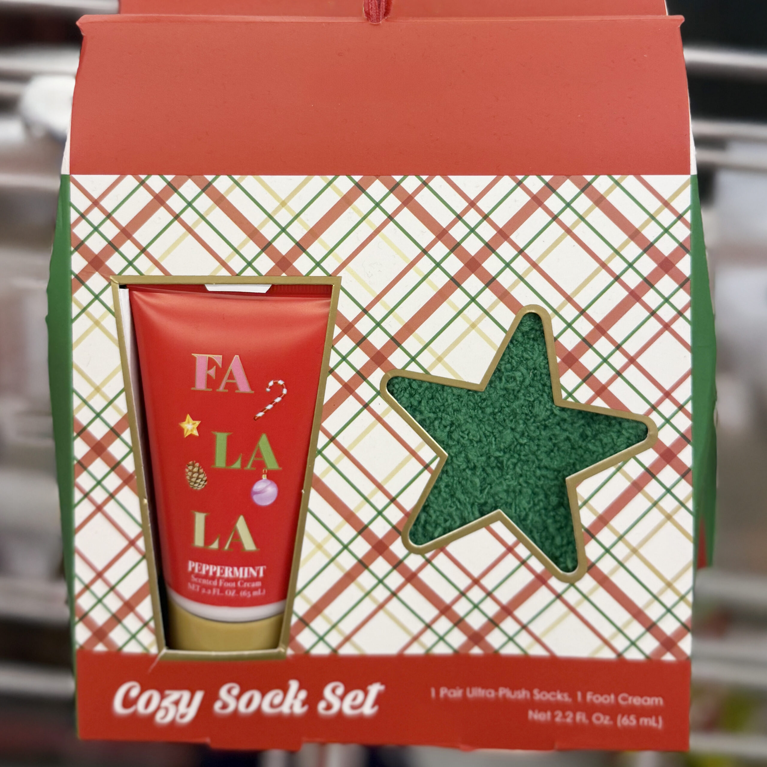 Holiday-themed cozy sock set in a red and green plaid box. The packaging features a red tube of peppermint foot cream labeled “FA LA LA” with festive icons and a star-shaped cutout revealing green plush socks. Text at the bottom reads “Cozy Sock Set.”